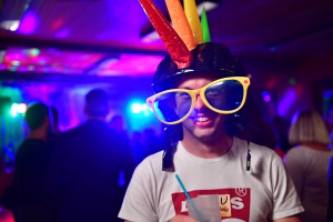 Lächelnder Mann mit gelben Partybrillen und Partyhut, im farbigen Licht einer Feier; Details sollen Feststimmung vermitteln.