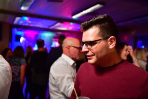Junger Mann mit Brille steht bei einer Feier im Vordergrund, blickt nach unten und hält einen Drink; bunte Lichtkulisse im Hintergrund.