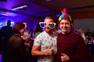 Mehrere Feiernde in einer beleuchteten Partyhalle, im Vordergrund drei Personen mit lustigen Partyaccessoires, lächelnd und in Gespräche vertieft.