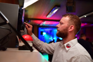 Mann bedient Computer-Display mit der Hand an einem Messe-/Festplatz; im Hintergrund bunte Bühnenbeleuchtung.