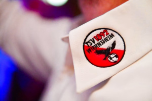 Weißes Stoffpatch mit rundem Aufnäher und roter/ schwarzer Grafik, auf Schuh-Zunge angenäht; dient als sichtbares Abzeichen.