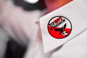 Weiße Stofffläche mit rundem Patch: rotes Feld, schwarze Figur und Schriftzug, als dekoratives Abzeichen auf Kleidung.