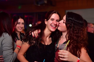 Drei lachende junge Frauen in einem festlichen Innenraum, eng beieinander, eine hält ein Getränk mit Strohhalm, Gesprächsstimmung.