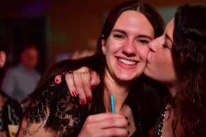Zwei lachende Frauen posieren eng beieinander, eine winkt mit der Hand ins Bild, im Hintergrund unscharfe Partybeleuchtung.