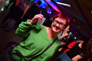 Lachende Frau im grünen Outfit im Festsaal, hält Getränkbecher hoch und winkt im Hintergrund bei farbigen Lichtern eine Menge.