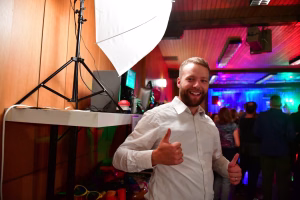 Lächelnder Mann mit zwei nach oben zeigenden Daumen vor Fotozubehör; große Softbox beleuchtet ihn, Hintergrund zeigt Partylicht.