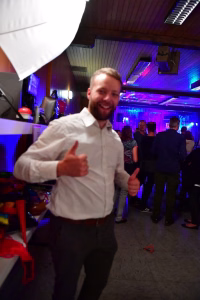 Lächelnder Mann im weißen Hemd auf einer Feier, Daumen nach oben in der Vordergrund-Szene, beleuchteter Raum mit Gästen im Hintergrund.