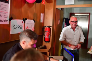 Mann lächelt an der Tür, neben Kasse-Schild; roter Feuerlöscher und Bedienungstisch im Hintergrund während einer Veranstaltung.