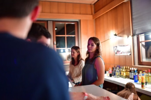 Zwei Personen an einer Bar, eine blickt nach vorn; im Hintergrund stehen Flaschen, warmes Licht während eines Events.