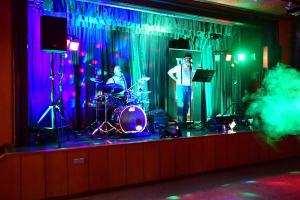 Bühne mit Schlagzeug und Sänger am Mikrofon, Scheinwerfer in Grün und Blau, Bühnenset für Live-Musik und Veranstaltung.
