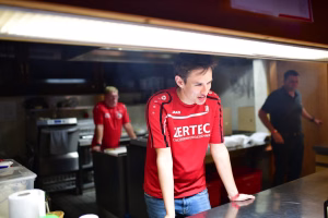 Mann in rotem Arbeitsshirt bewegt sich in einer Werkstatt vor Maschinen und Tischen, im Hintergrund Kollegen bei der Arbeit.