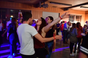 Tanzpaar in Partybeleuchtung auf einer Veranstaltung, Armhaltung beim Tanzen; im Hintergrund Publikum, Dekor und beleuchtete Bühne.