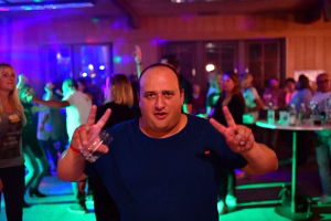 Mann im Vordergrund bei einer Feier im Scheinwerferlicht, fotografiert beim Zuhalten mit zwei erhobenen Fingern; Hintergrundgäste und Tische.
