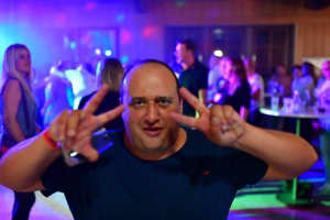 Feierabend-Partyraum mit blau-violetter Beleuchtung; eine Person im Vordergrund macht „Lass uns los“-Handgesten und deutet aufs Publikum.