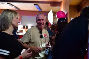 Feiernde Person stößt mit einem Glas an; mehrere Gäste im Hintergrund, Ballons und Partydeko im Innenraum als festliche Stimmung.