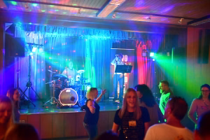 Musikgruppe auf einer Bühne mit Mikrofon, Schlagzeug und Lichtshow in Blau und Rot; Publikum tanzt im Vordergrund.