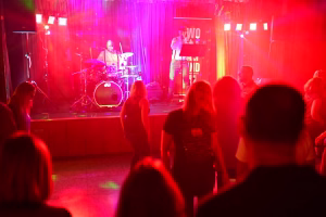 Rockband auf einer Bühne bei rotem Bühnenscheinwerferlicht; Publikum im Vordergrund, Performer mit Mikrofon und Schlagzeug im Einsatz.