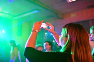 Besucherin fotografiert mit Smartphone ein Bühnenkonzert bei buntem Licht; Blick auf Bildschirmspiegelung, Musiker im Hintergrund.