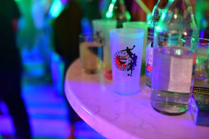 Party-Getränkebecher mit Vereinslogo auf einer beleuchteten Tafel, im Hintergrund weitere Gläser und Flaschen.