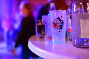 Plastikbecher mit Vereinswappen und Fußballmotiv auf einer Festtafel, unscharfer Hintergrund mit Partylicht und Personen.