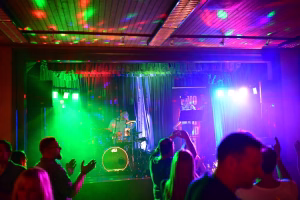 Musikbühne mit Schlagzeug, Vorhang und bunten Scheinwerfern; Publikum im Vordergrund, beleuchtetes Konzert-Setup für eine Live-Performance.