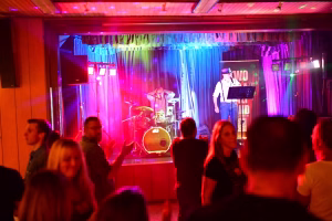 Bühne mit Schlagzeug und Musiker am Mikrofon; farbige Lichtstimmung, Vorhang und Publikum im Vordergrund als Konzert.
