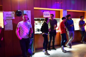 Gesellige Gruppe bei einer Bar, im Hintergrund Schilder wie Pfand-Rückgabe und Kasse, im Vordergrund Gespräche mit Getränken.