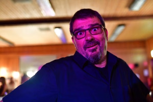 Porträt einer Person mit Brille, behelmter Gesichtshaltung und freundlichem Lächeln bei Veranstaltung in warmem Licht.