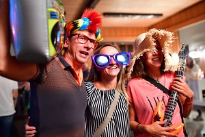 Feiernde Menschen mit bunten Partyhüten und Sonnenbrillen; eine Person hält eine Deko-Gitarre, lachend in einer Innenumgebung.