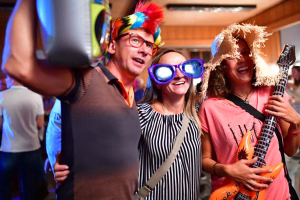 Feiernde Menschen mit Partyhut und Sonnenbrille, halten ein Instrument und schauen in die Kamera; ausgelassene Stimmung im Innenraum.