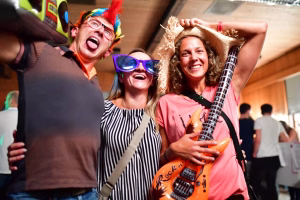 Drei Personen feiern mit Partyhüten und Accessoires, eine Person in der Mitte jubelt, rechts hält jemand eine Gitarre; ausgelassene Stimmung.
