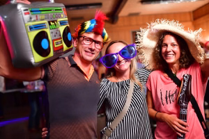 Drei Personen feiern mit Partybrille und Federhut im Innenraum; sie halten Mobilteil/Fotoapparat und ein Spielzeuginstrument, lachend.