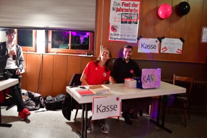 Zwei Personen sitzen an einem Tisch mit Kasse-Schild, neben einem Ausschank; im Hintergrund Flyerwand und Luftballons als Eventdekoration.
