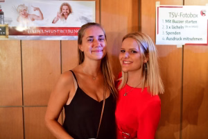Zwei Frauen lächeln nebeneinander vor einer Wand, im Hintergrund ein Schild mit Schrittfolge zur Fotobox-Aktion.