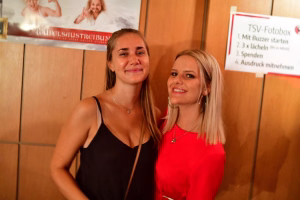 Zwei lächelnde Frauen im Innenraum bei einer Veranstaltung, vorne mit Blick in die Kamera; Hintergrund mit Hinweisschilden.