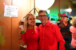 Zwei Personen im roten Outfit posieren lächelnd, im Hintergrund ein Banner mit Fotowettbewerb und farbiges Partylicht zur Feier.