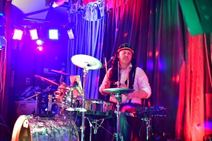 Perkussionist am Schlagzeug mit Mikrofon, umgeben von farbigem Bühnenlicht und Vorhang—Aktionsaufnahme während einer Live-Feier.