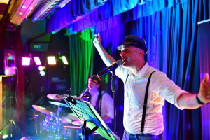 Musiker mit Hut am Mikrofon, Arm in Richtung Publikum; farbige Bühnenlichter und Vorhang im Hintergrund, begleitet von weiteren Instrumenten.