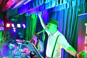 Sänger mit Hut am Mikrofon, Arm erhoben, Bühnenperformance mit farbigen Lichtern und Vorhängen im Hintergrund.