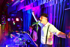 Sänger mit Hut am Mikrofon, Arm erhoben, während Band und Lichtshow im Hintergrund; Bühnenauftritt bei Festveranstaltung.