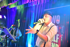 Sänger im Scheinwerferlicht mit Mikrofon und Handgesten auf einer Bühne, im Hintergrund Vorhang und Musikerinstrumente zur Live-Show.