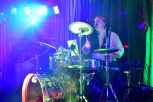 Musiker singt am Mikrofon mit Drumset und Synth-Pads, Bühnenlicht in Blau und Grün zeigt Unterhaltung bei einer Festveranstaltung.