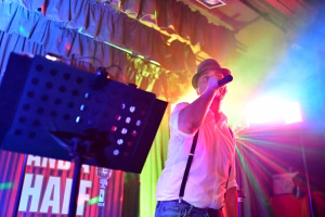 Sänger mit Hut und Mikrofon auf einer Bühne, beleuchtet von bunten Lichtstrahlen, mit Projektions-Backdrop und Lautsprecher im Vordergrund.