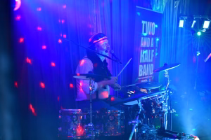 Musiker am Mikrofon, spielt Schlagzeug unter blauem Bühnenlicht; im Hintergrund eine beleuchtete Bühne, geeignet für Live-Performance.