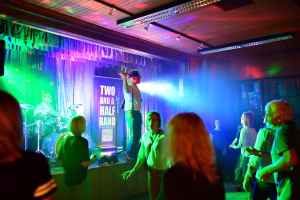 Musikabend in der Halle: Sänger mit Mikrofon auf der Bühne, Band im Hintergrund, Publikum im Vordergrund, buntes Bühnenlicht und Nebel erzeugen Stimmung.