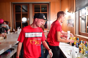 Mehrere Personen in roten Shirts arbeiten an einer Bar, mischen Getränke und servieren Gäste in einem beleuchteten Innenraum.