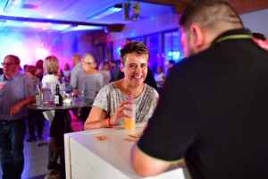 Fröhliche Person am Tisch hält ein Glas mit Strohhalm und winkt lachend; im Hintergrund Gäste bei festlicher Beleuchtung.