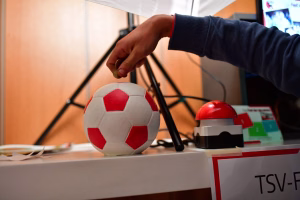 Hand hält eine Tischtennisplatte, roter Sportball liegt bereit neben Spielgeräten; symbolisiert ein Vereins- bzw. Sporttreffen.