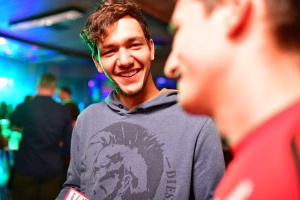 Lächelnder Mann in Partybeleuchtung in einer Feierstätte, im Vordergrund eine unscharfe Person; freundliche Stimmung.