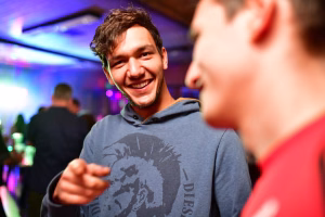 Lächelnder Mann im Partyraum, im Vordergrund redet/gestikuliert er in angeregter Unterhaltung; unscharfe Personen im Hintergrund, Lichtstimmung.